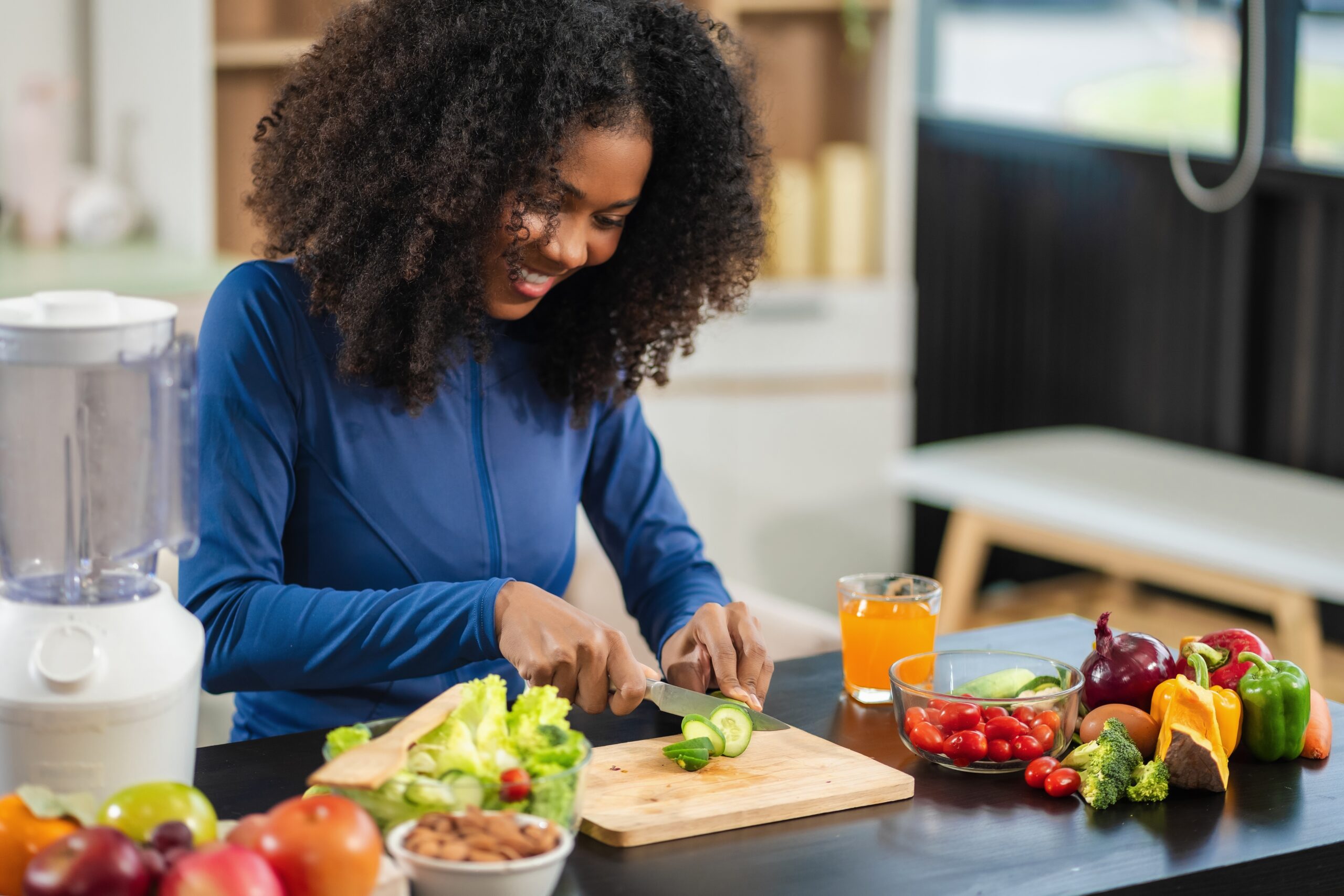 Black women standing at the counter cutting a cucumber with other vegetables laid out on the counter top, practicing healthy living
